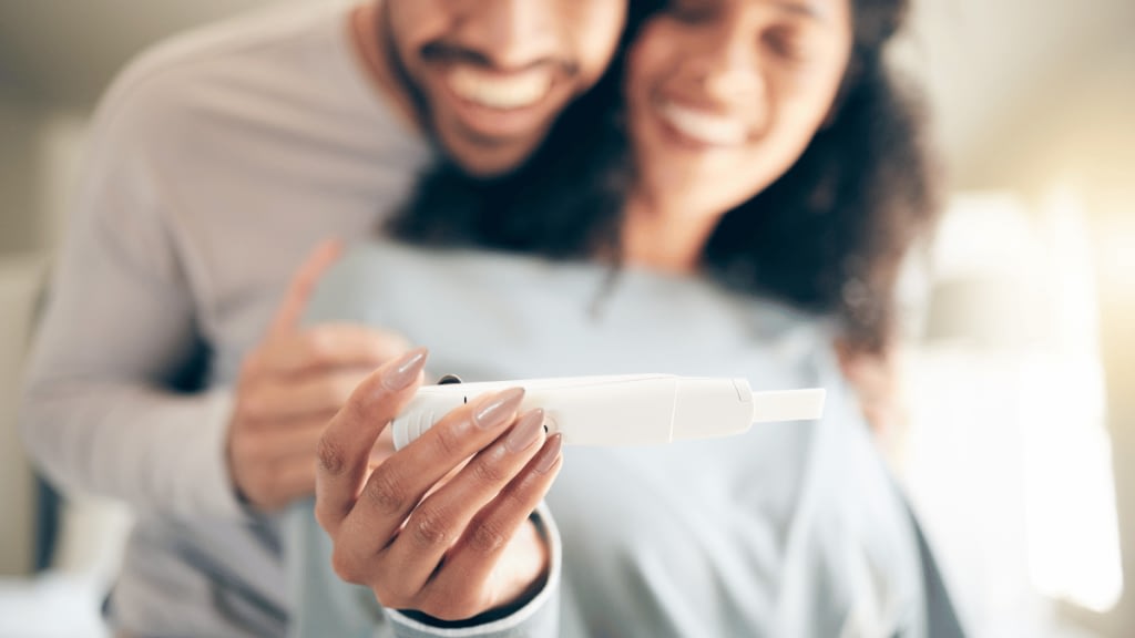 Smiling Mixed race lesbian woman showing ultrasound beside caucasian pregnant girlfriend waving hand at laptop sitting on sofa in living room at home