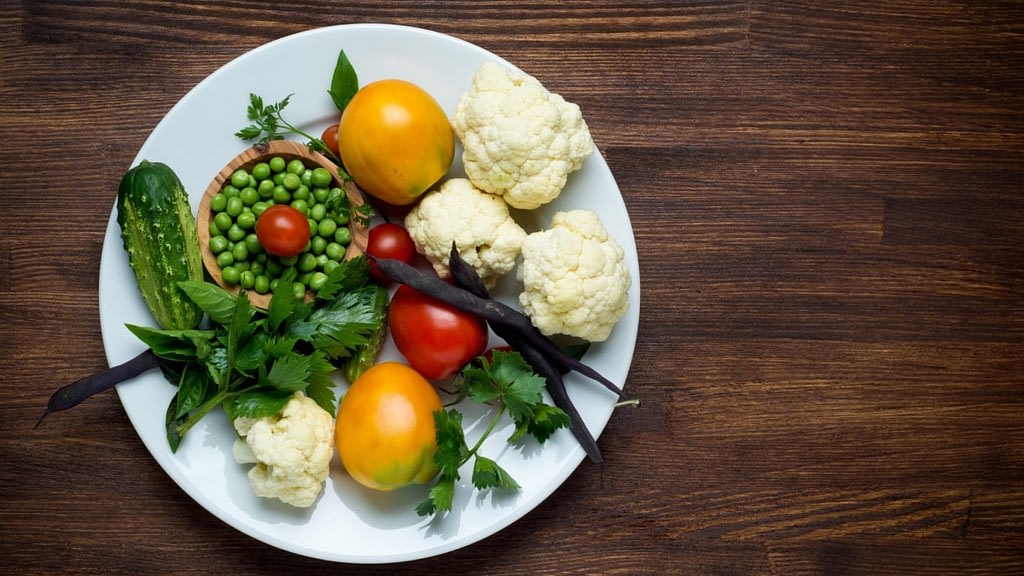 A photograph of a plate of food with tomatoes, cauliflower, peas, a cucumber and fresh herbs.