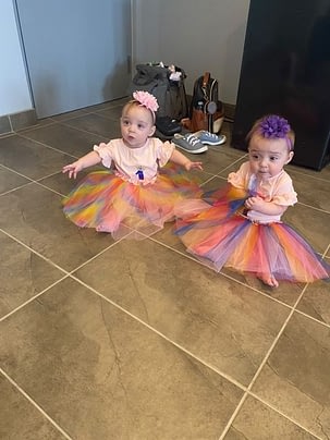 A set of twins sit on the floor wearing colourful tutu's and bows in their hair