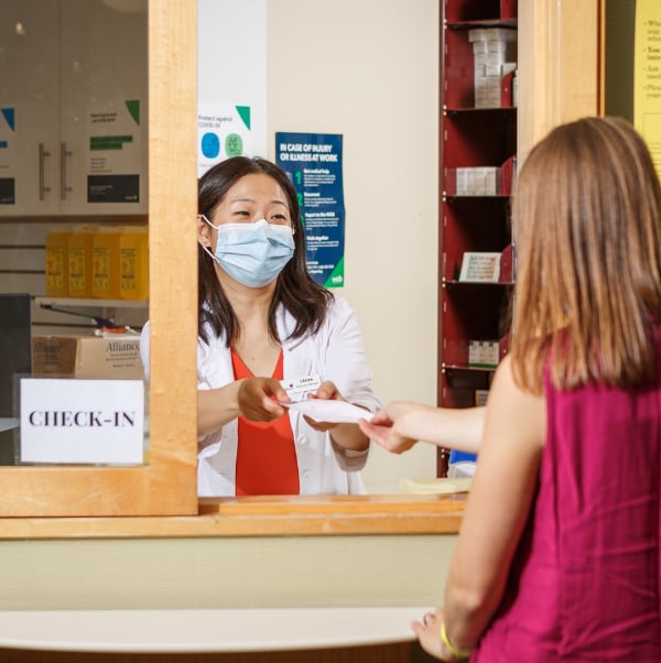 A photograph of a pharmacist wearing a mask handing a prescription to a patient.