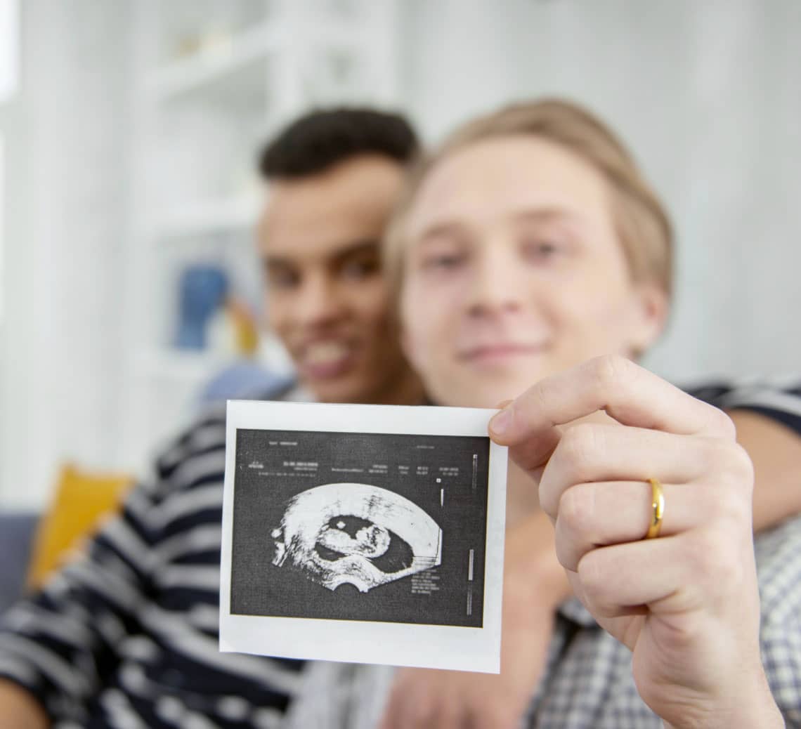 A photograph of two men hugging and holding an ultrasound photo.