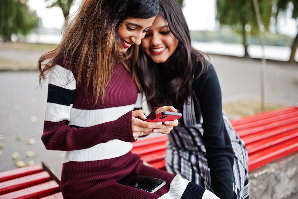 Two smiling people sitting on a bench and looking at a smartphone one of them is holding.