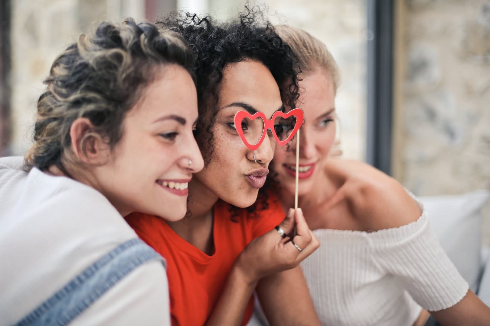 Three people sitting with their heads near each other and all looking in the same direction. The middle person holds costume heart shaped glasses in front of their eyes.
