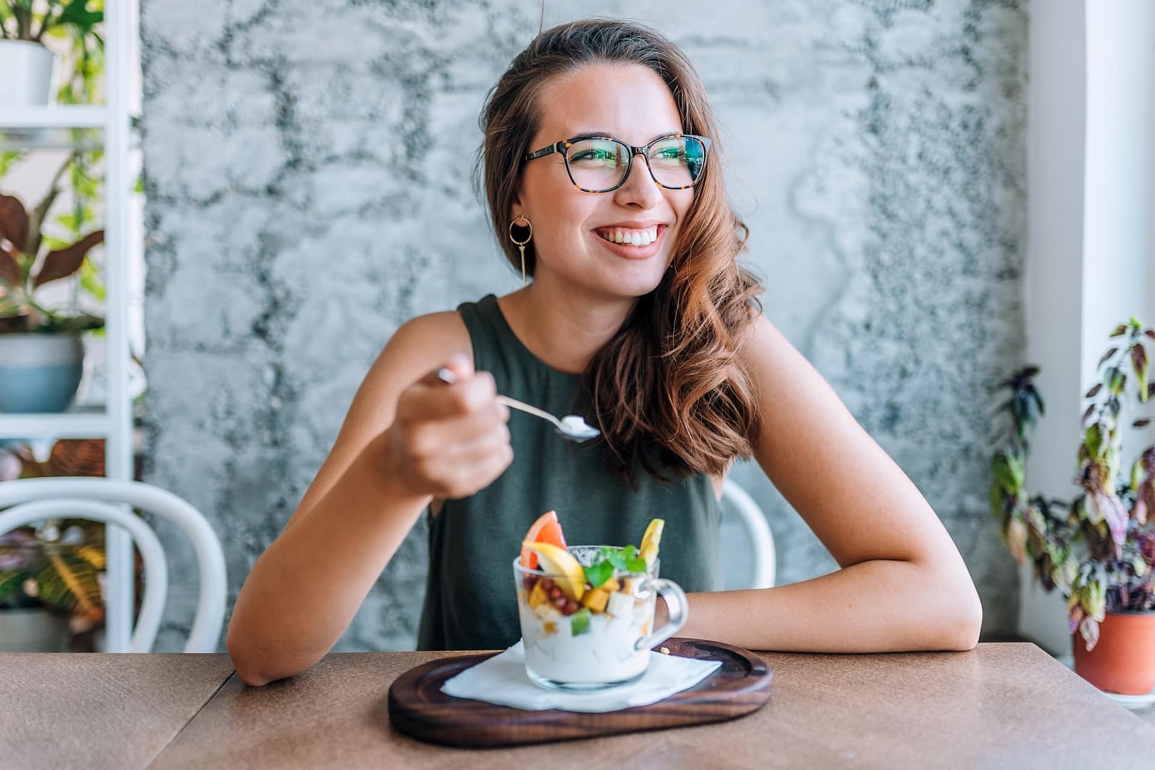 A smiling person sitting at a table eating from a clear mug containing fruit.