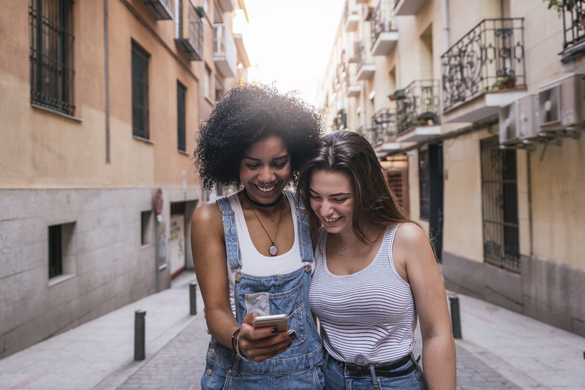 Two smiling people with their arms around each other both looking at a smartphone one of them is holding.