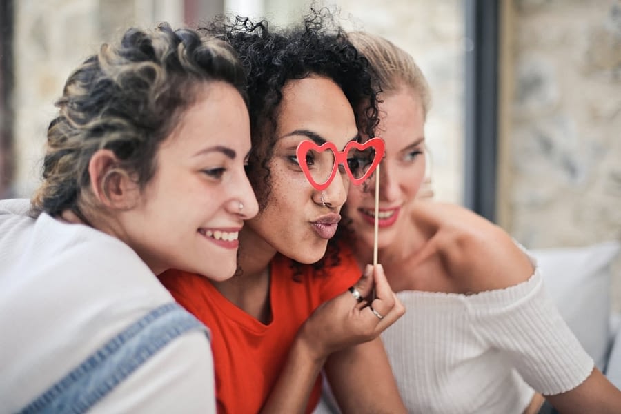 Three people sitting with their heads near each other and all looking in the same direction. The middle person holds costume heart shaped glasses in front of their eyes.