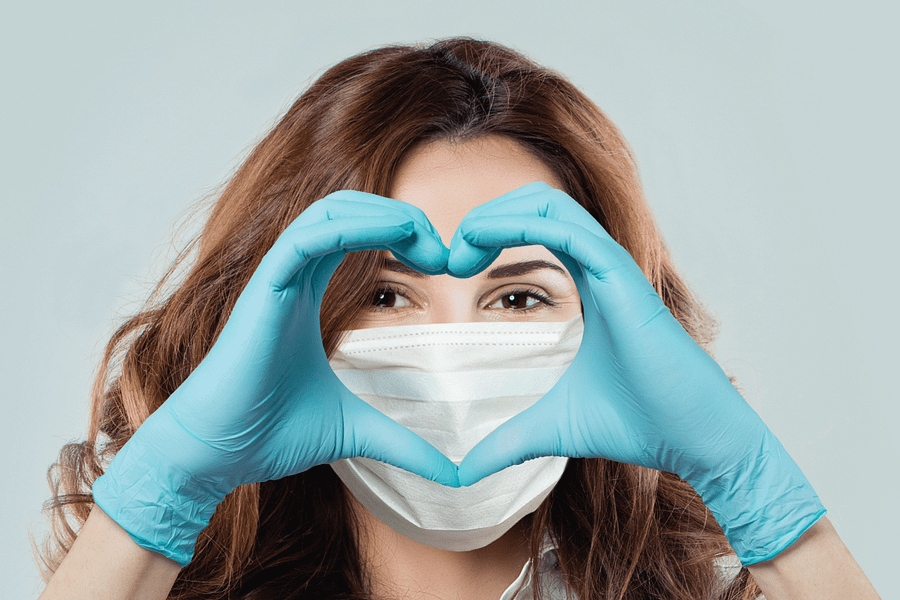 A doctor wearing a P P E mask holds their gloved hands in the shape of a heart in front of their face.